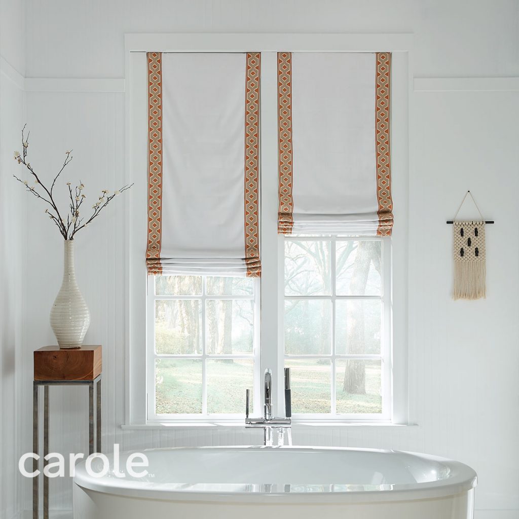 A white tub in a white bathroom with Flat Roman Shades on the windows in white with vertical trimming in a paprika and beige geometric pattern.