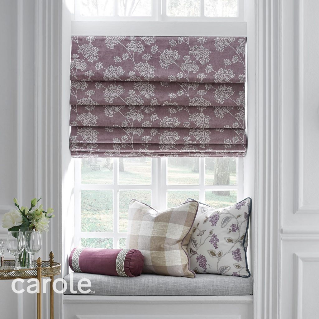 A pillow covered window seat in a white room with a Hobbled Roman Shade in mauve and a delicate white floral pattern on the window.