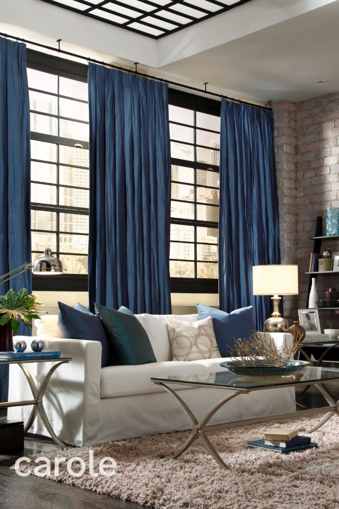 Blue Tuxedo Pleat Drapery over a wall of windows in a brick building with an off-white sofa and light beige shag rug.