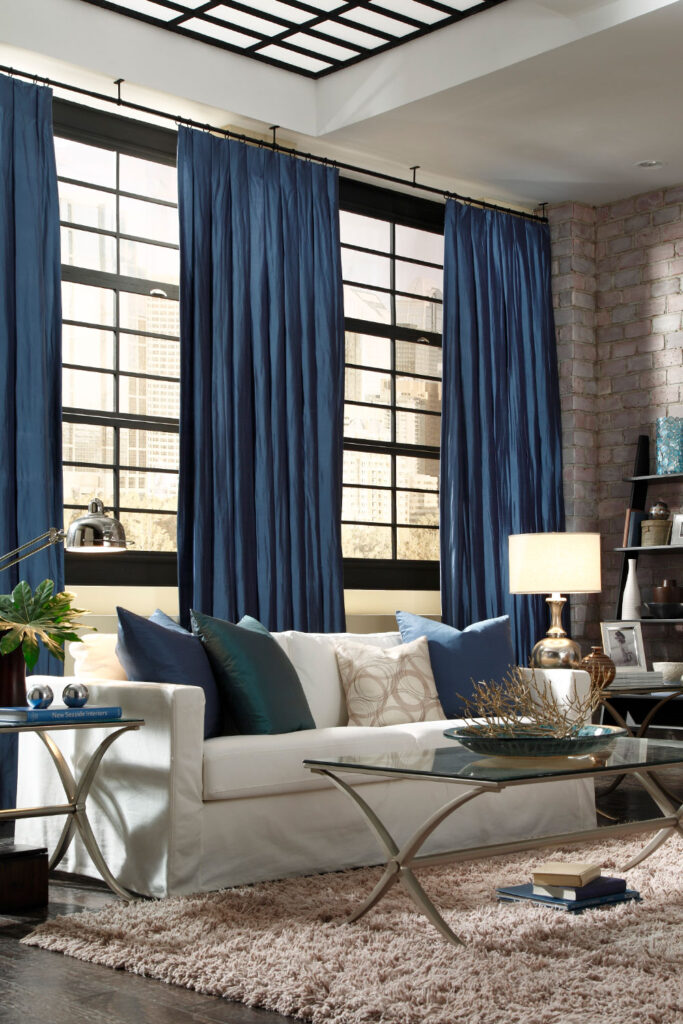 Blue Tuxedo Pleat Drapery over a wall of windows in a brick building with an off-white sofa and light beige shag rug.