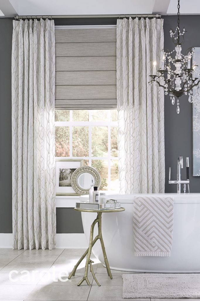 A white tub and ornate crystal chandelier in a gray and white room with lightly patterned white Two Finger Pinch Pleat Drapery over light grey Front Fold Roman Shades.
