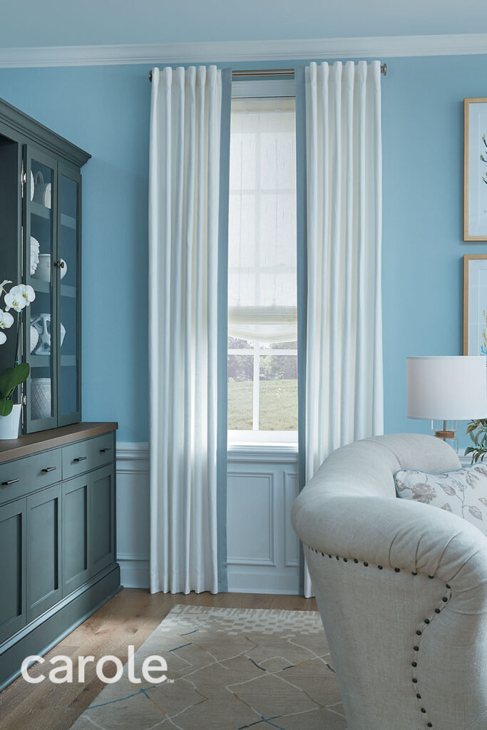 Floor to ceiling white Hidden Tab Top Drapery with light blue banding on the leading edge over a white Sheer Soft Roman Shade on a window in a light blue living room with light neutral and dark green decor.