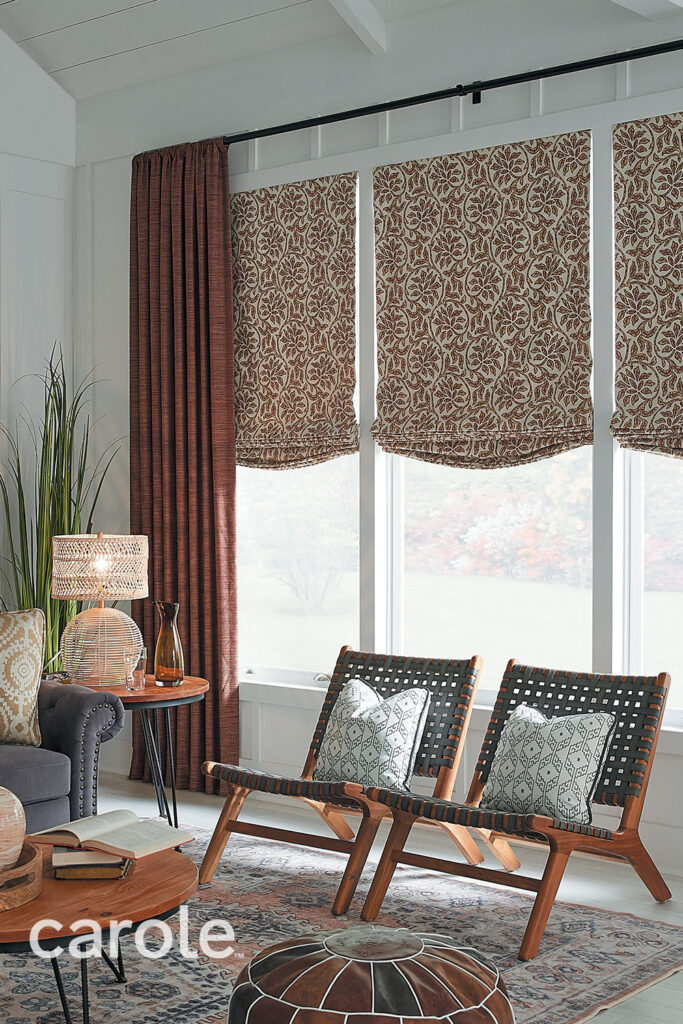 A casual living room with a wall of three windows dressed in subtly textured brick red Rod Pocket Drapery and black drapery hardware with French returns over Soft Roman Shades in a large scale two-toned geometric floral.