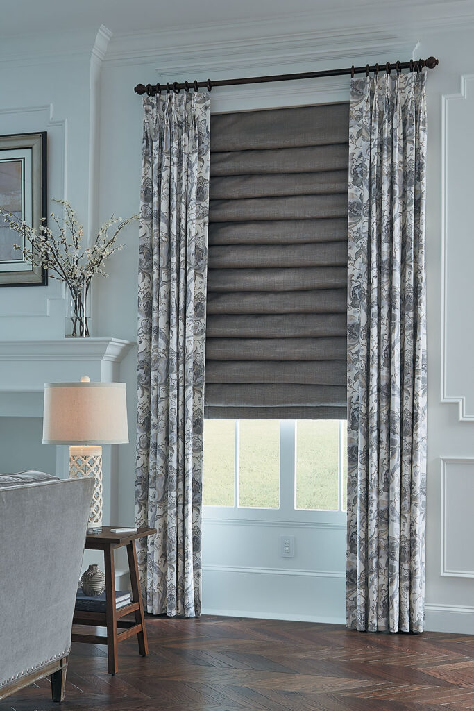 A window in a living room with Three Finger Pinch Pleat Drapery in a large scale neutral colored floral and a Hobbled Roman Shade in brown fabric with a subtle sheen.