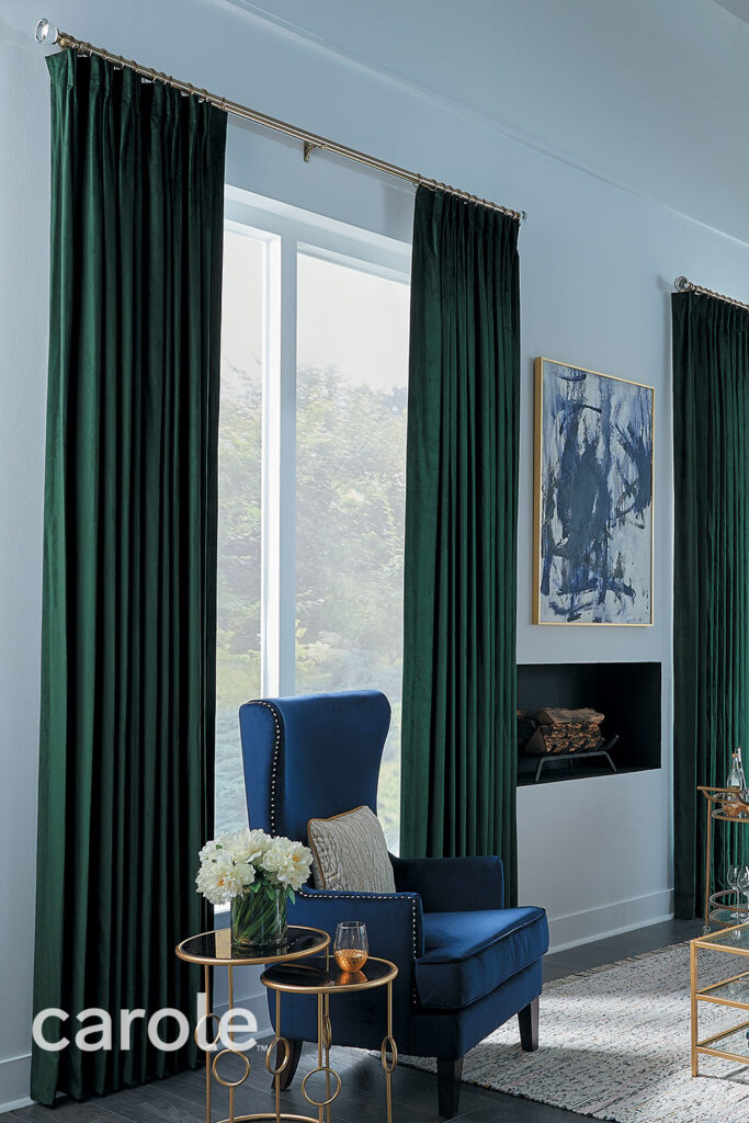 Two Finger Pinch Pleat Drapery in dark green velvet with gold drapery hardware and glass finials in a living room with a dark blue velvet chair and accent tables in gold and glass.