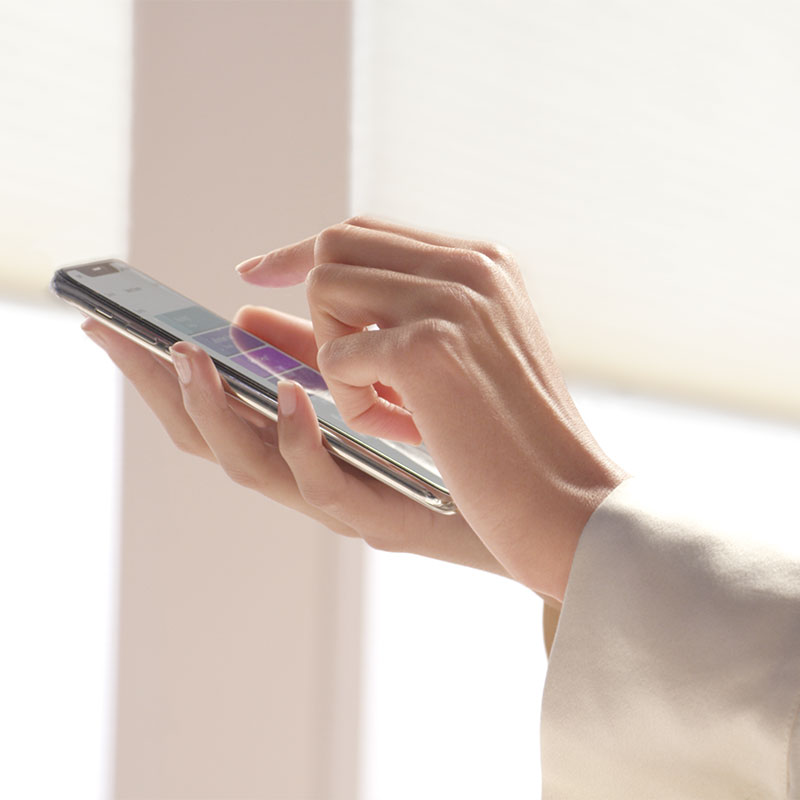 A woman's hands as she uses AMP motorization on a smart phone with window shades in the background.
