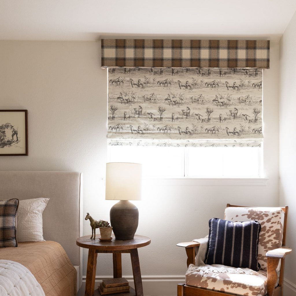 A bedroom in brown and natural colors featuring a window with a cornice in a large scale plaid fabric and a partially closed soft shade with at pattern of playful horse line drawings.