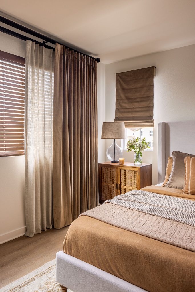 A bedroom in warm brown tones featuring two layers of drapery over blinds on one window and a soft Roman shade on another window.