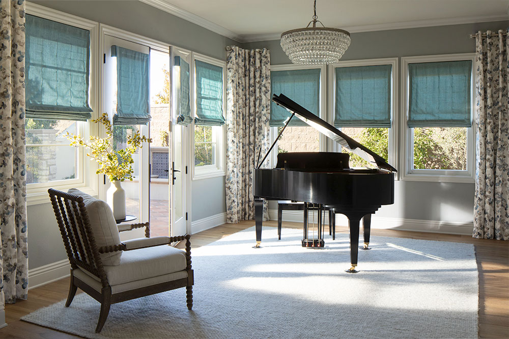 A sunroom featuring a grand piano with teal soft Roman Shades on all of the windows and drapery panels at the corners of the room.