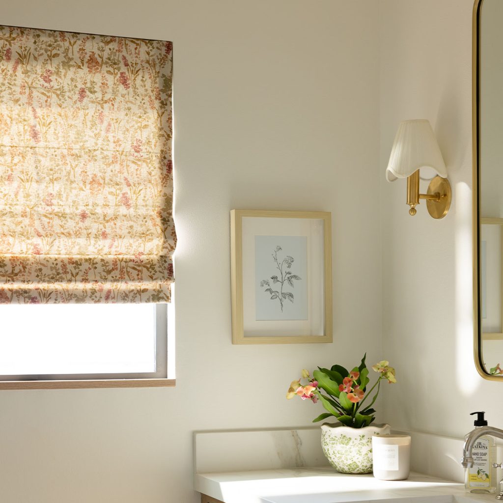 Detail of a window next to a bathroom sink, featuring a partially opened Flat Roman Shade in a delicate rose and neutral floral pattern.