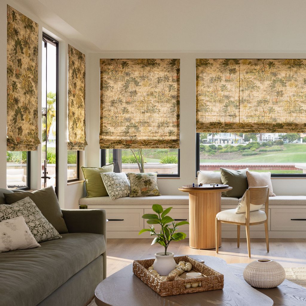 A comfortable family room in deep greens and light neutrals with a sofa, bench seating and small table and chair, featuring two walls of windows with Flat Roman Shades in a mid-tone neutral foliage pattern.