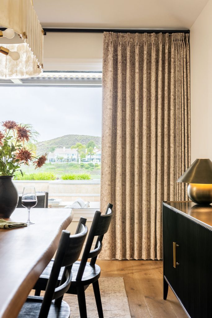 A dining room in browns and blacks featuring a large picture window with subtly patterned neutral drapery on black drapery hardware.