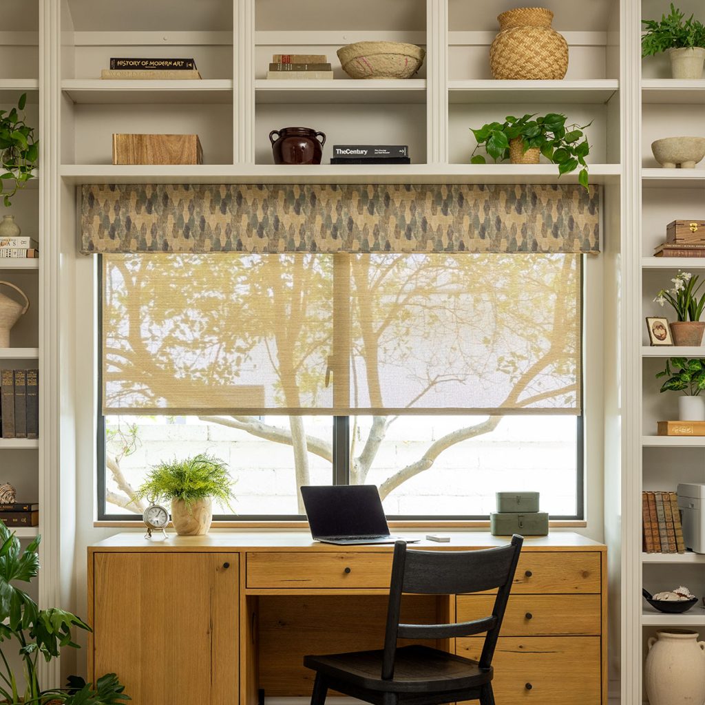 An office with built-in floor-to-ceiling booskshelves featuring a desk under a large window. The window is adorned with a Low Profile Statler Cornice in an earth-toned elongated hexagon pattern over a semi-sheer shade.