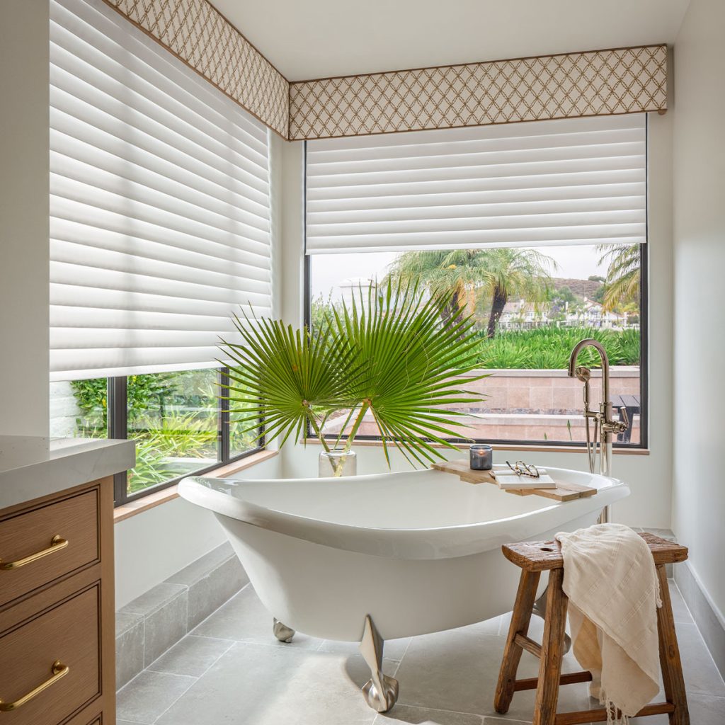 A bathroom featuring a claw-foot bathtub surrounded by large picture windows on two sides. The windows have Statler Cornices meeting in the corner in a brown and ivory rounded diamond pattern over opaque ivory colored shades.