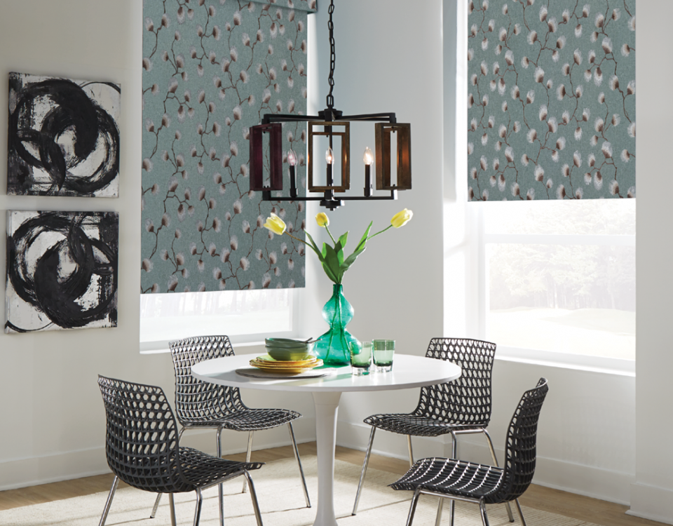 Small dining area with windows that have fabric roller shades in a floral embroidery pattern from Carole Fabrics.